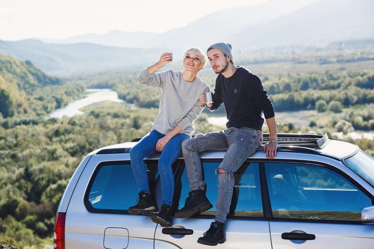 A couple of young people are sitting on the roof of a car against the backdrop of a mountain valley