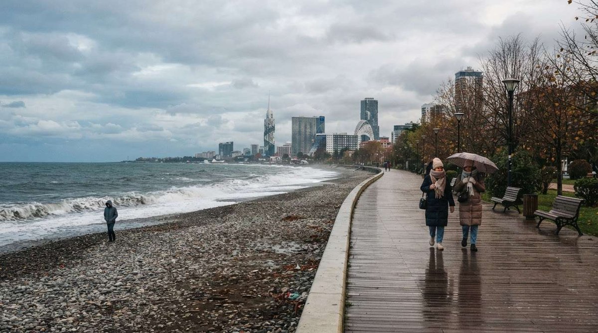 People walking along Batumi's promenade with an umbrella as waves crash on the pebble beach, with the city skyline visible in the distance.