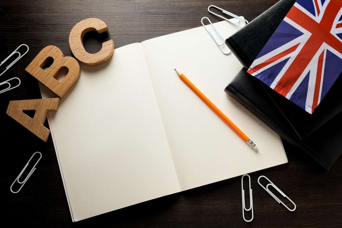 An open blank notebook with a pencil sits beside wooden ABC letters, paperclips, and a small British flag on a dark wooden surface.