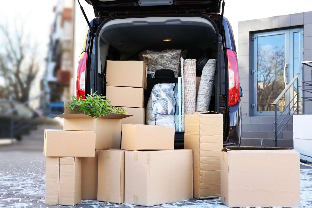 A car’s open trunk and the pavement around it filled with cardboard boxes, a rolled rug, and a houseplant outside an apartment building.