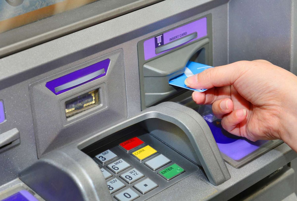 A close-up of a hand inserting a blue card into an ATM's card reader, with the numeric keypad and cancel, correction, and enter buttons below.