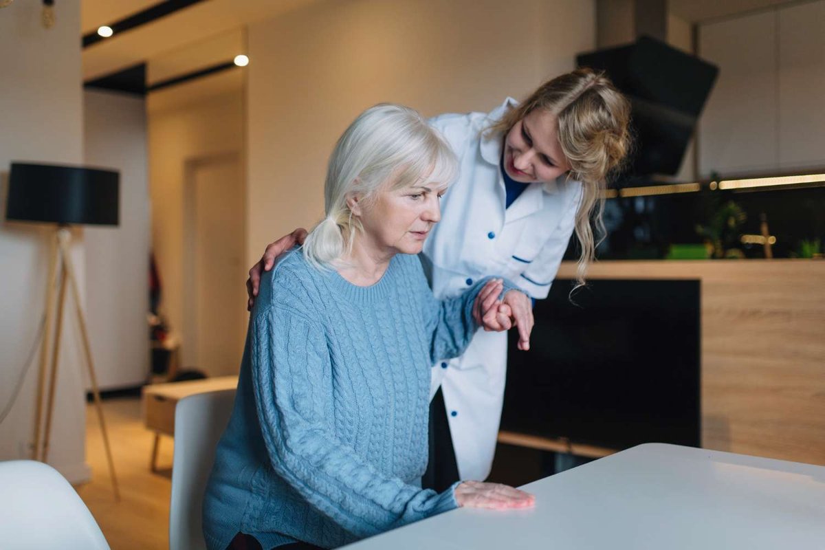 A woman in a white coat places a hand on an elderly woman’s shoulder while helping her at a kitchen table.
