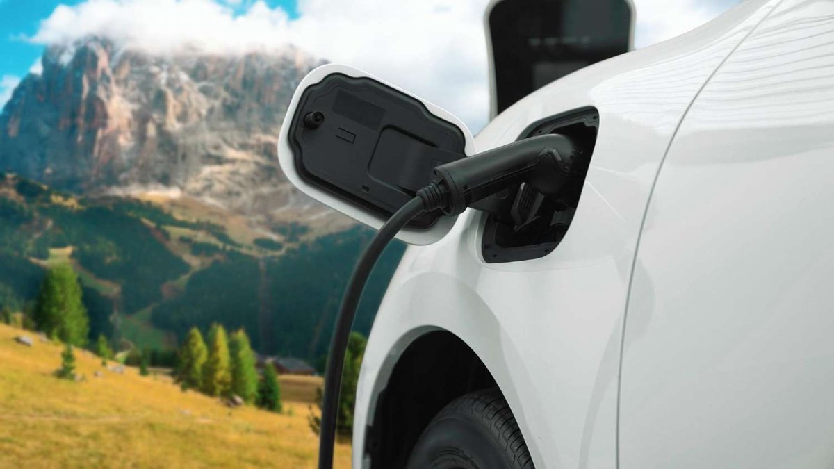 A close-up of a charging connector inserted into a white car’s port, with a rocky mountain range and alpine meadow in the background.