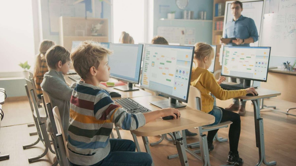 Children at desktop computers working with a visual block-coding program while a teacher instructs in the background — illustrating programming classes for kids in Tbilisi, Georgia.