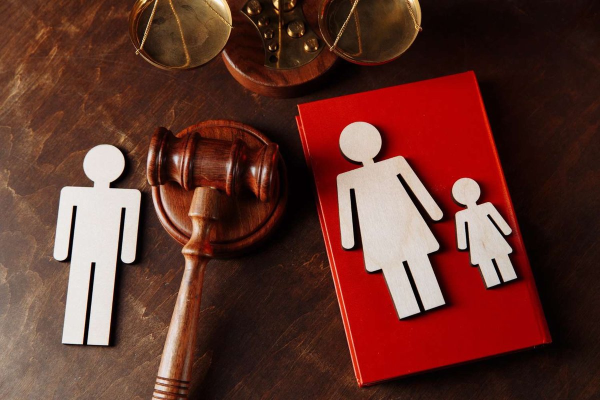 Wooden cutout figures of a separated man, mother, and child stand beside a gavel, scales of justice, and a red law book on a wooden table.
