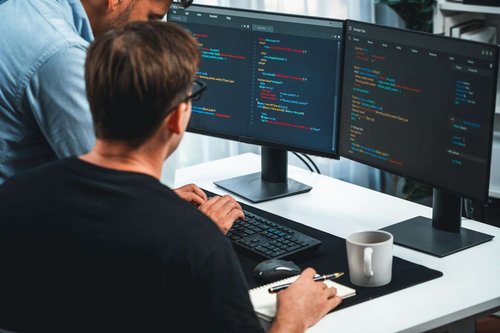 Two men reviewing PHP code across dual monitors at a desk with a notebook and coffee mug — illustrating computer and programming courses in Tbilisi, Georgia.
