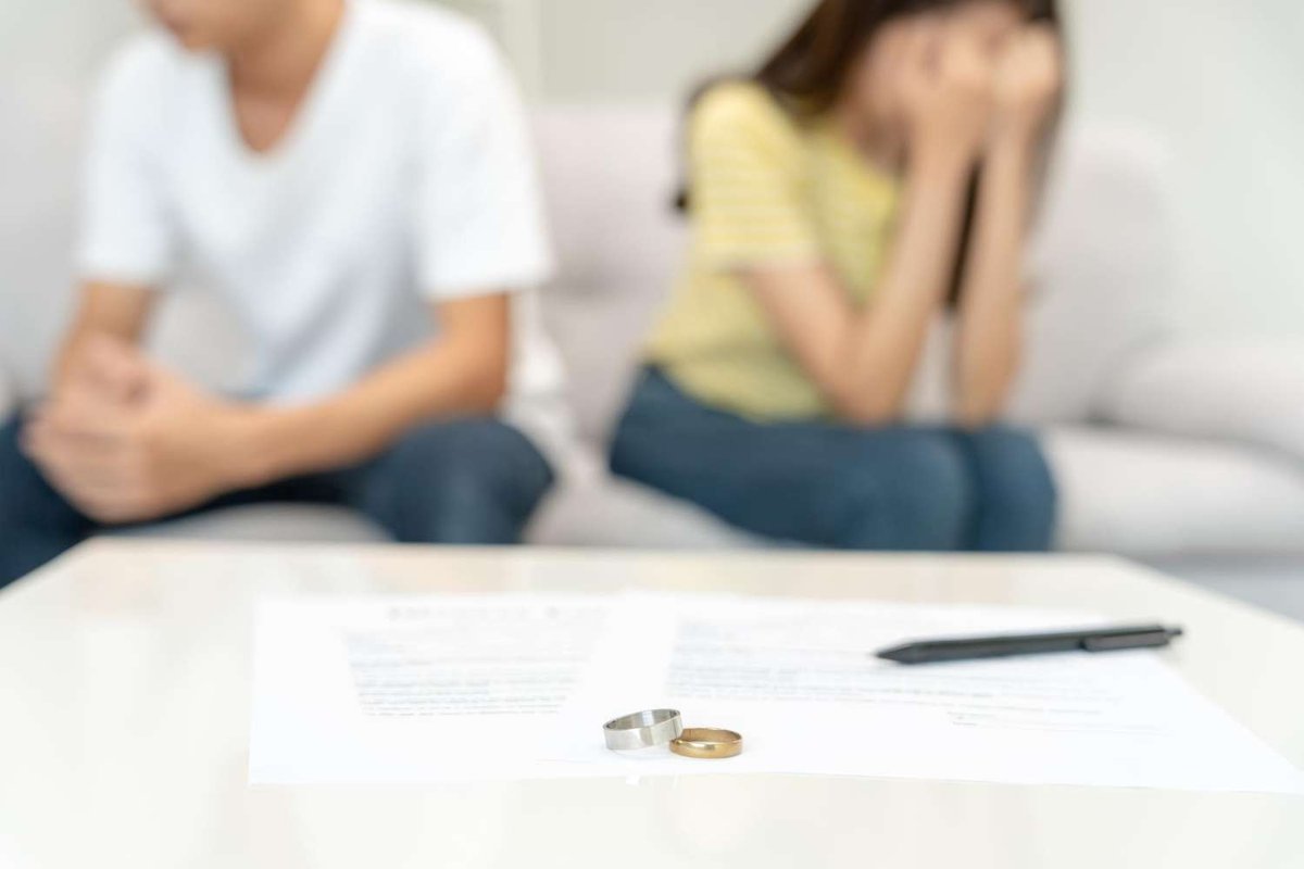 Two wedding rings rest on a signed divorce document beside a pen, with an upset couple sitting apart on a couch in the background.
