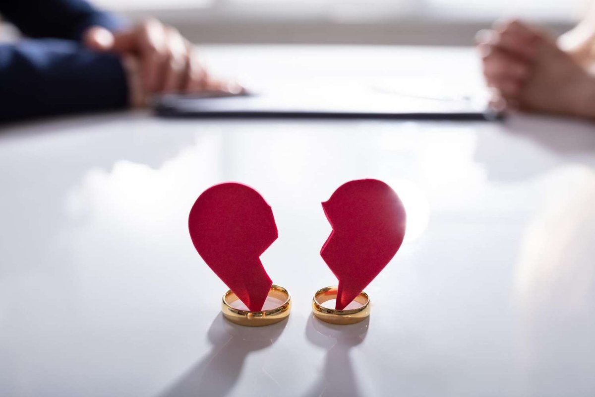 Two gold wedding rings each hold half of a torn red paper heart on a table, with a man and woman seated across from each other in the background.