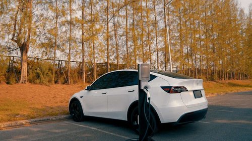 A white electric SUV parked at a curbside charging post with its cable connected, surrounded by golden autumn trees at sunset.