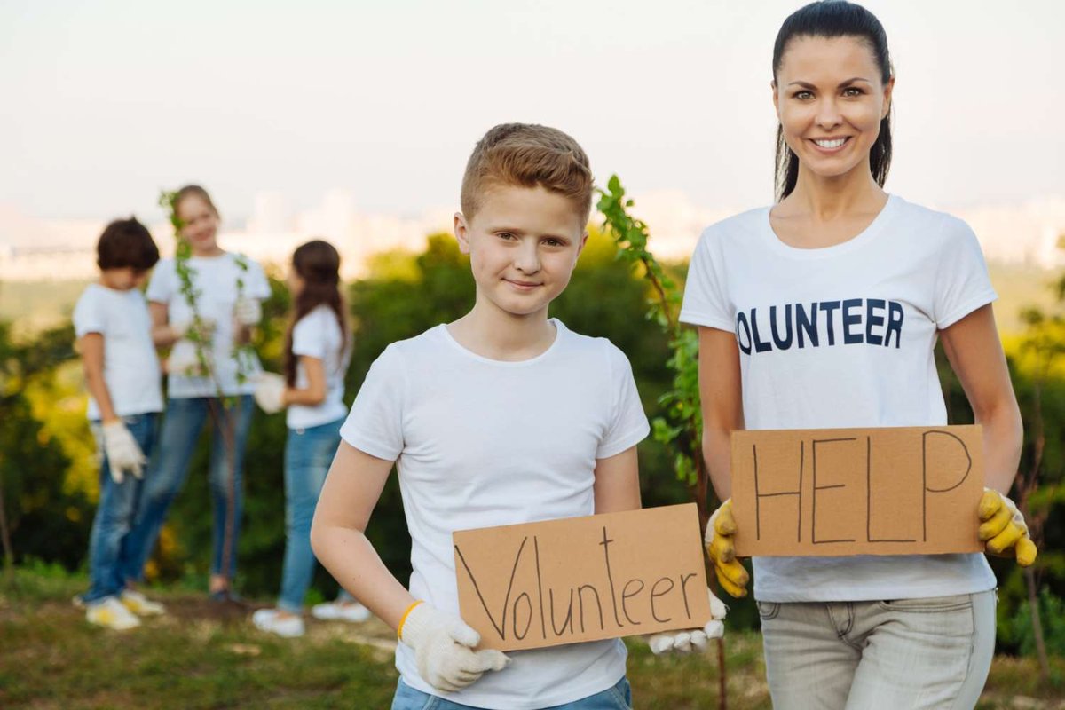 A boy and a woman wearing a volunteer t-shirt hold hand-lettered cardboard signs reading “Volunteer” and “Help,” with children planting a tree sapling in the background.
