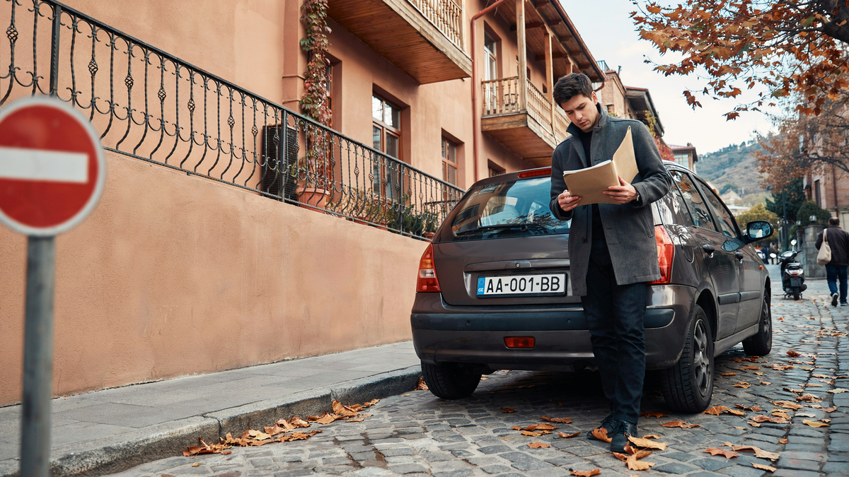A man reviews his vehicle documents beside a Georgian-plated car on an old Tbilisi street, marking the year-from-entry rule that governs how long a foreign driver's license stays valid.