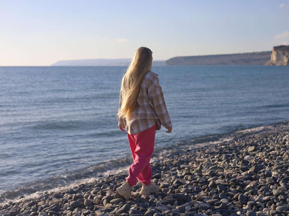 A woman walks the quiet pebble shore in warm layers as coastal cliffs rise in the distance, marking the tail end of swimming season.