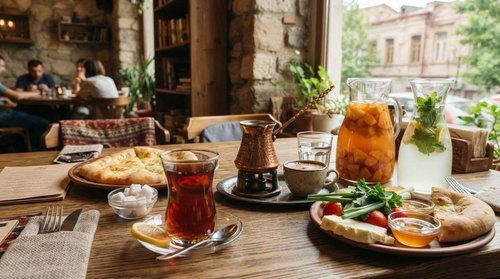 A café table set with black tea, Turkish coffee in a copper cezve, fruit compote, and mint lemonade, alongside puri bread and a cheese plate by a window.