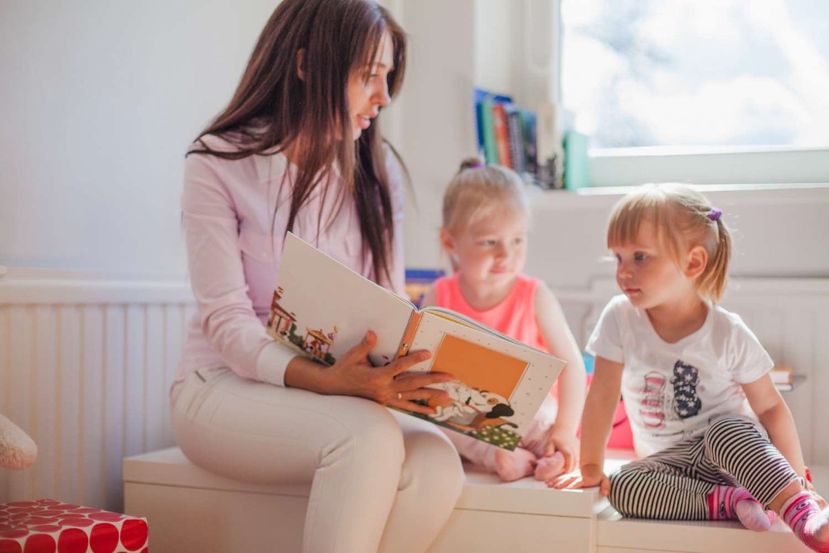 A governess reads an illustrated picture book while two young girls sit beside her on a bench in a bright playroom with bookshelves by the window.