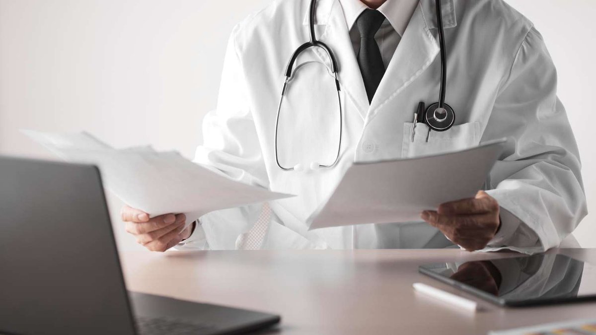 A doctor in a white coat reviews paperwork at his desk, illustrating the medical documentation involved in choosing health insurance coverage.