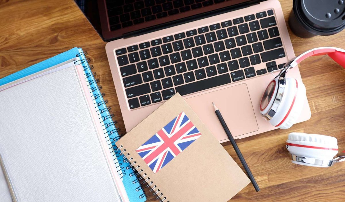 A flat lay shows a laptop keyboard, a notebook with a Union Jack sticker, a pencil, headphones, and a coffee cup on a wooden desk.