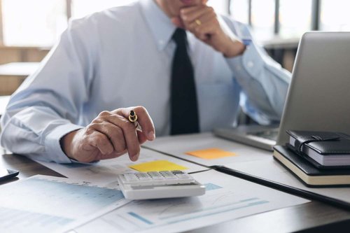 A man in a shirt and tie thinking while holding a pen near a calculator and printed charts, with a laptop and notebooks on the desk.