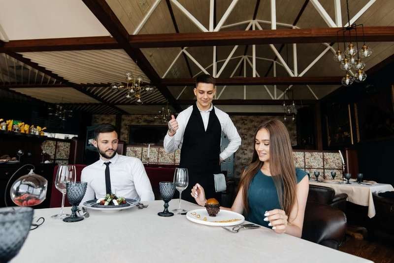 A young waiter in a stylish apron serves a table with a beautiful couple in an elegant restaurant