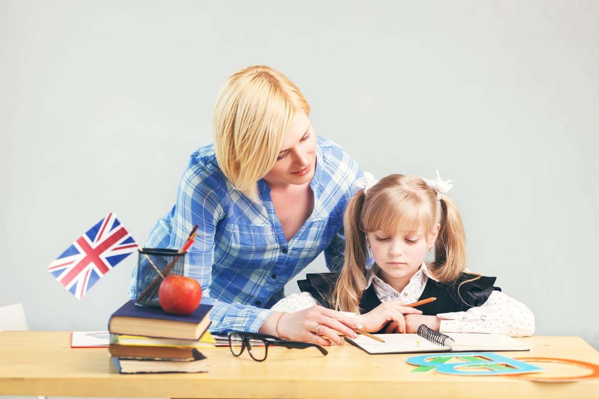 A woman in pigtail braids writes in a notebook while a woman leans over to help her, with stacked books, an apple, and a small British flag on the desk.
