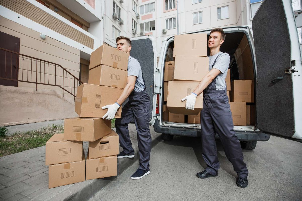 Two gloved movers in overalls load stacked cardboard boxes into a white van parked outside a residential apartment building.