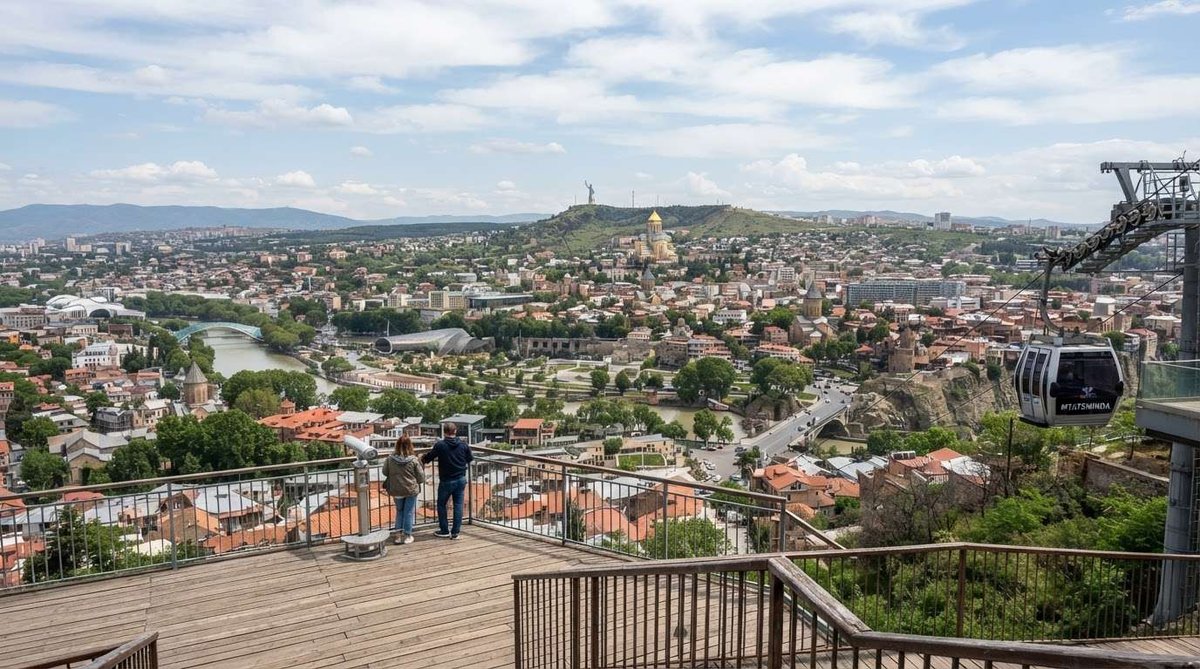 Visitors at a railing viewpoint over Tbilisi's old town, river, and bridges, with a Mtatsminda cable car gondola passing on the right.