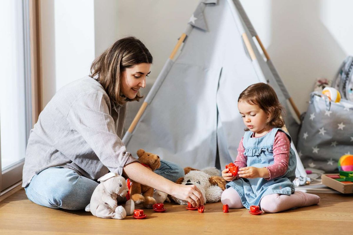 A nanny and a young girl sitting on the floor playing with a toy tea set and stuffed animals in front of a play tent.