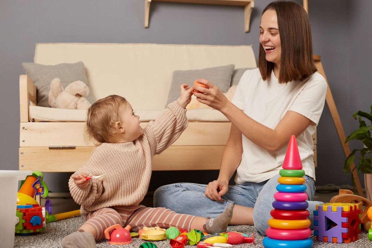 A nanny laughs while playing with a toddler surrounded by a stacking ring toy, blocks, and stuffed animals on a living room floor.