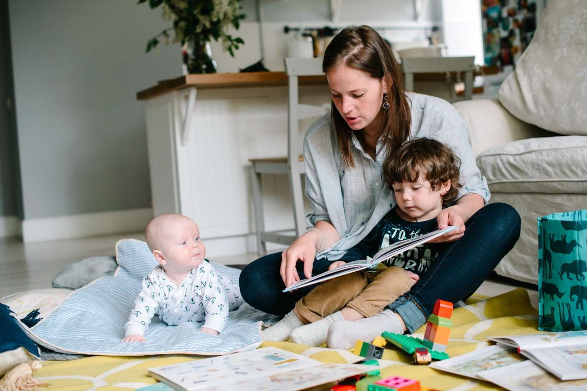A nanny reads a book to a toddler sitting on her lap while a baby looks on from a play mat, surrounded by building blocks and toys on a living room floor.