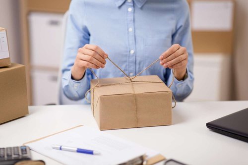 A woman tying twine around a brown paper-wrapped package at an office desk, with shipping boxes, a laptop, and paperwork nearby.