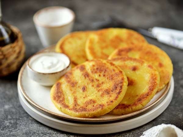 A stack of golden cornbread rounds sits on a plate next to a small bowl of sour cream.