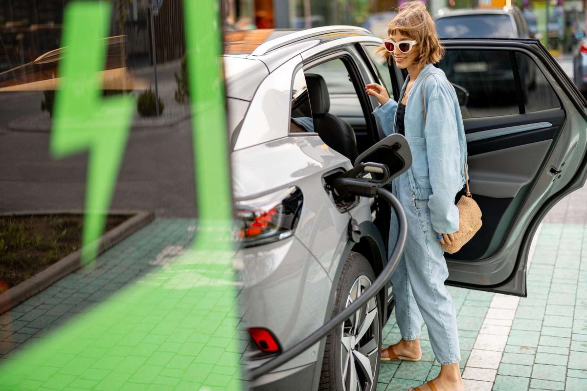 A woman in a denim outfit stands beside her car as its charging cable connects to the front port, with a green lightning-bolt sign in the foreground.