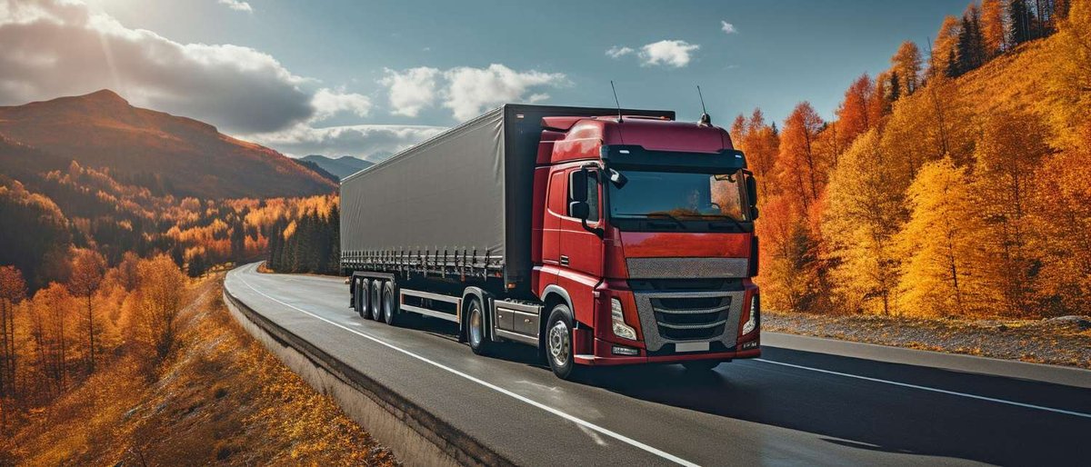 A red cabover truck with a curtain-side trailer driving along a winding road through hills covered in autumn foliage.