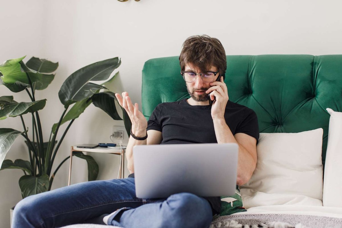 A man sits cross-legged on a green velvet sofa, laptop on his lap, talking on the phone with a concerned expression, surrounded by houseplants.