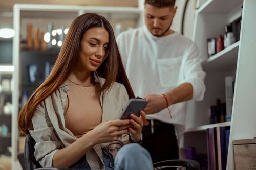 A happy woman writes a review on her smartphone while getting her hair styled at a beauty salon.