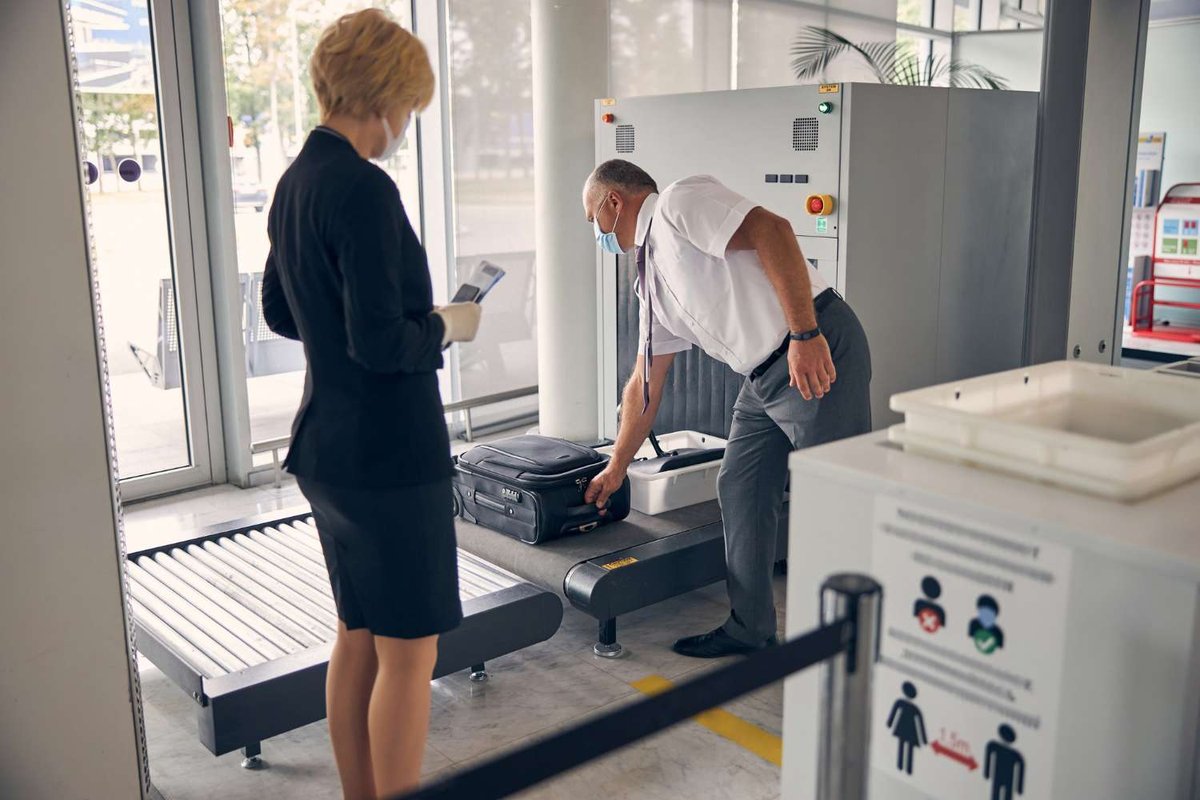 A security officer in a mask examines a suitcase on an X-ray scanner belt while an airline employee holding a boarding pass looks on.