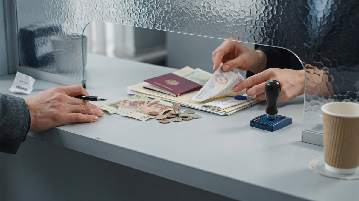 Hands at a service window handling a passport, papers, banknotes, coins, a pen, and a rubber stamp.