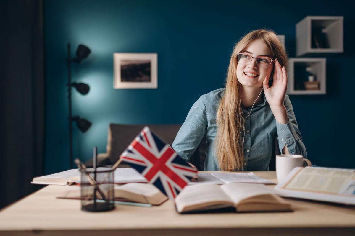 A young woman with glasses and an earphone smiles at her desk, surrounded by open books, a small British flag, and a coffee mug.