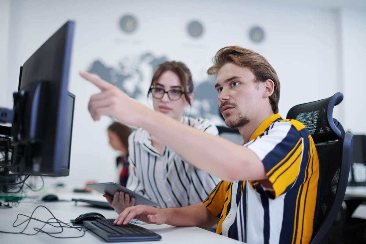 A man pointing at a screen while a woman holding a tablet looks on, seated at desks in a classroom-style office — illustrating a call for reviews on programming courses in Tbilisi, Georgia.