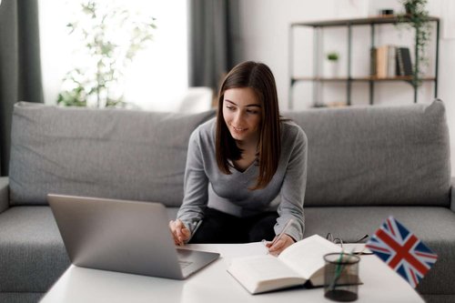 A woman sits on a couch at a coffee table, typing on a laptop beside an open notebook, pen, glasses, and a small British flag.