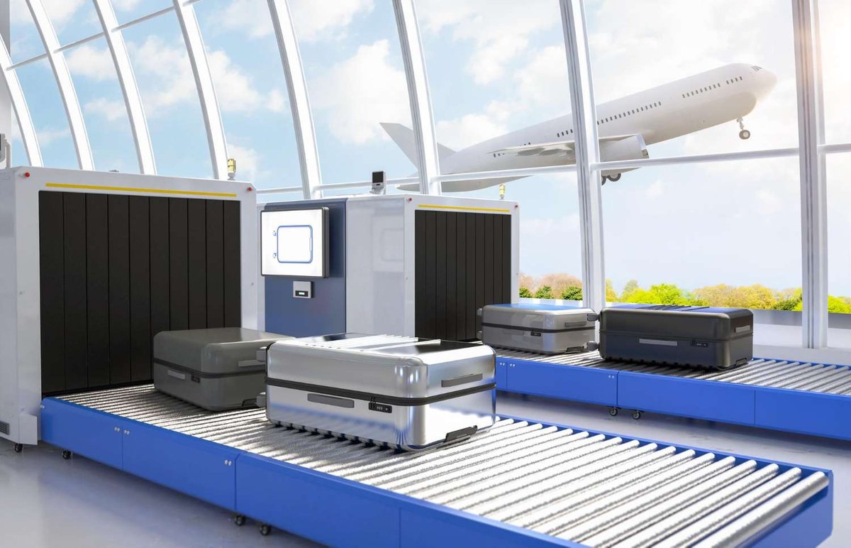 Luggage moving through an X-ray baggage scanner on blue conveyor rollers, with a plane taking off visible through the terminal windows.