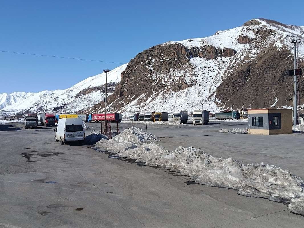 Trucks lined up in a snowy lot beneath towering, snow-capped mountains near the Kazbegi border crossing.