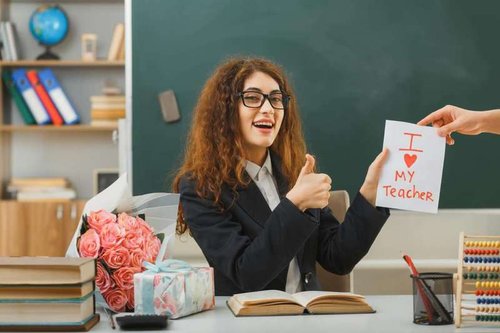 Smiling and giving a thumbs up, a young teacher received a card while sitting at her desk with school supplies in the classroom.