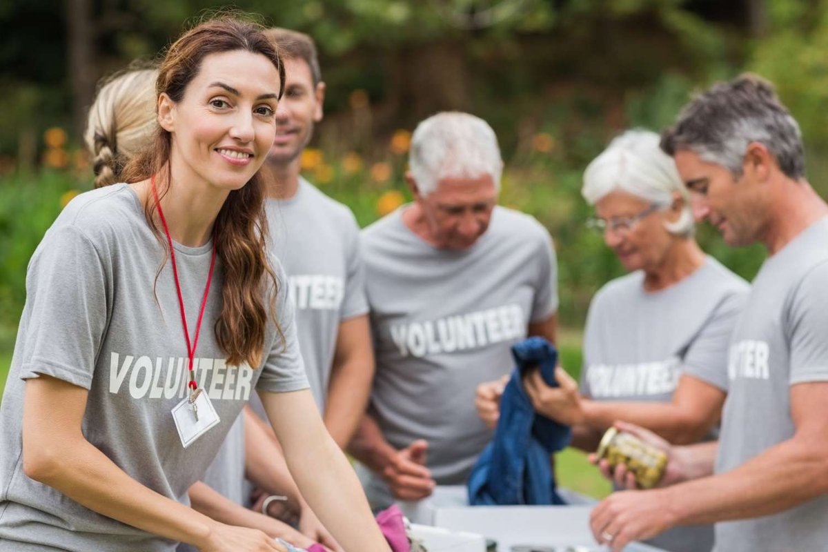 A smiling woman wearing a volunteer t-shirt and lanyard works alongside other volunteers sorting clothing and canned goods outdoors.