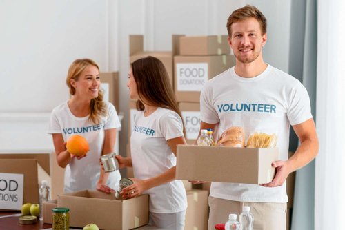 A man holds a box filled with bread, pasta, and water while two women behind him sort canned goods among stacked boxes labeled food donations.