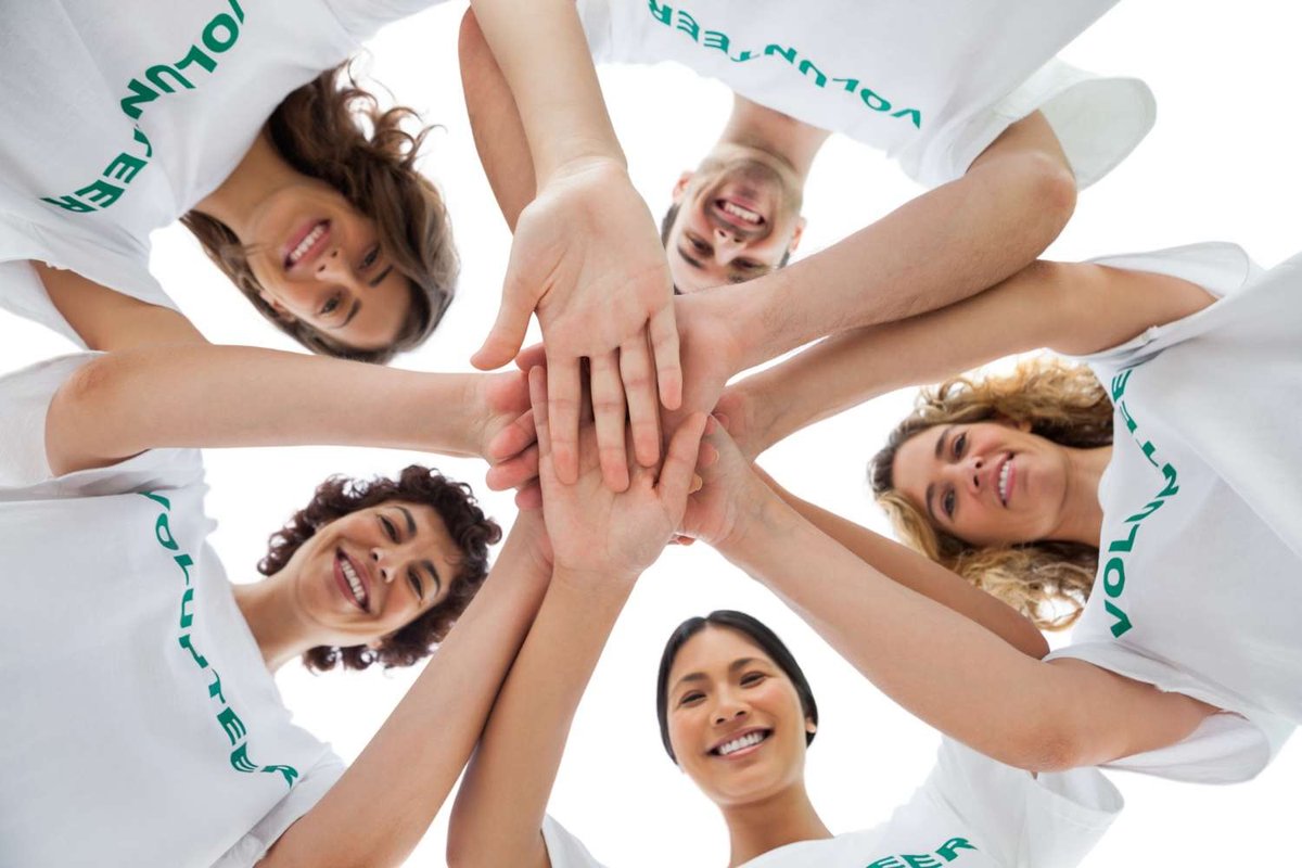 A group of smiling young volunteers in matching t-shirts stack their hands together, photographed from below against a bright sky.
