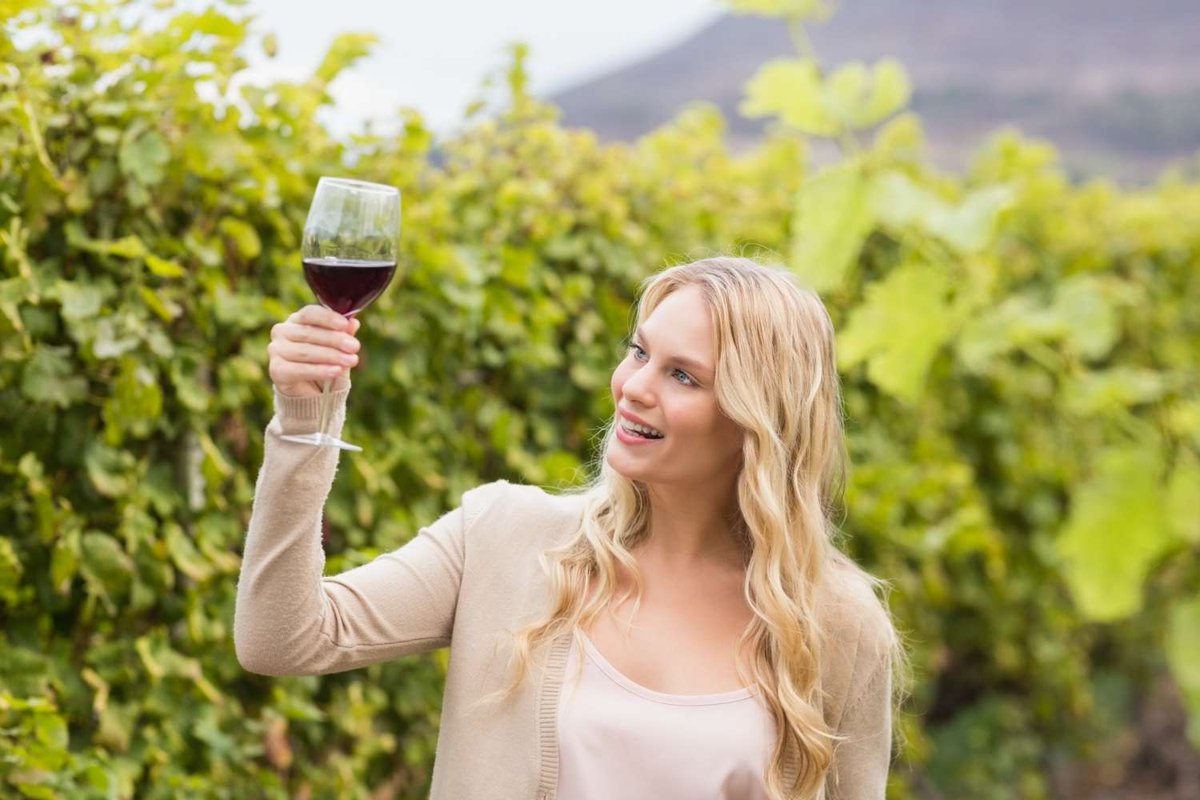 A smiling woman holds up a glass of red wine to examine its color against the green vineyard rows behind her.