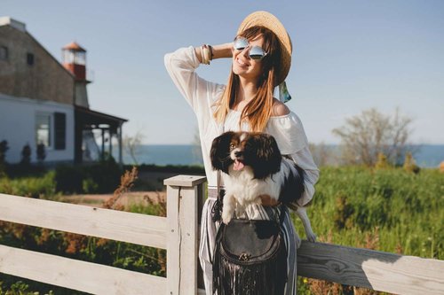 A woman in a straw hat holding her small dog beside a wooden fence, with a lighthouse and sea in the background — illustrating pet-friendly accommodation in Georgia.