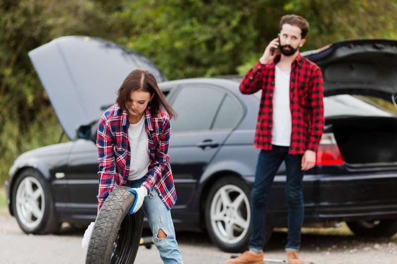 A woman holds a spare tire and a man talks on the phone and calls a tire shop.