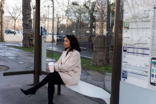 A woman sits at a public transport stop and holds a glass of takeaway coffee in her hands.