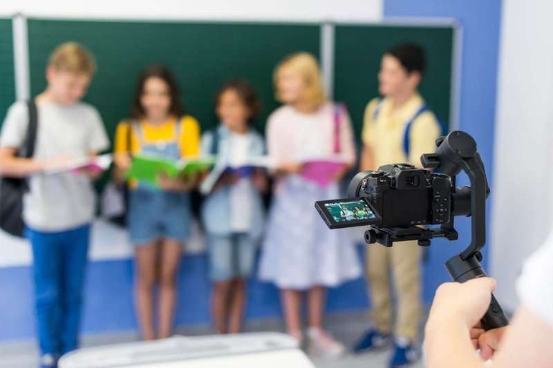 a group of children at a school blackboard with books in their hands, with a video camera pointed at them